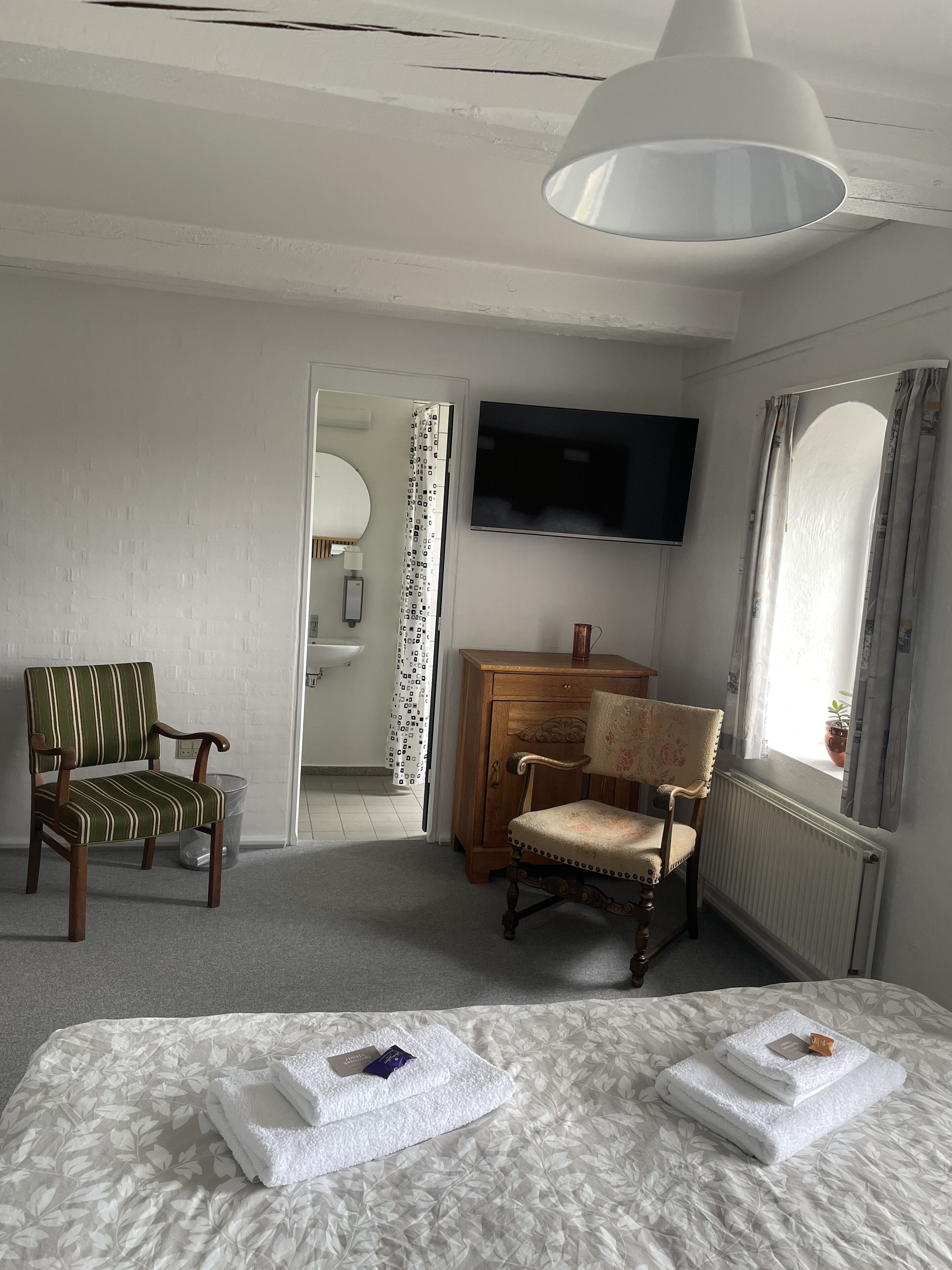 Ein Hotelzimmer mit Bett, zwei Sessel, Fernseher, Heizkörper und einem Fenster mit Vorhängen.