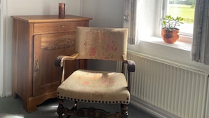 Une pièce avec un meuble en bois, un téléviseur fixé au mur, une chaise recouverte d’un tissu à motifs floraux et une fenêtre donnant sur un paysage verdoyant.