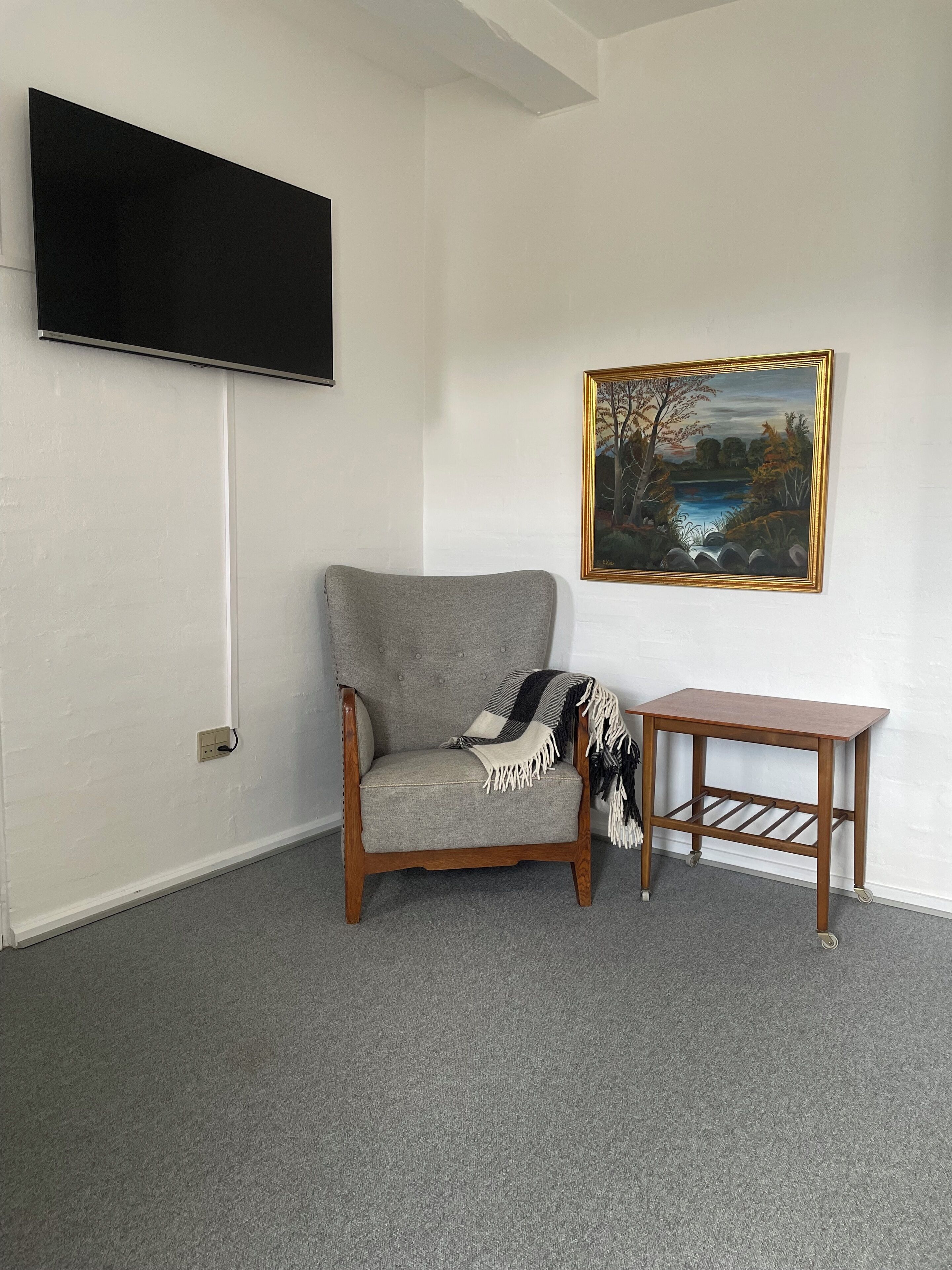 A room with a grey armchair, a small wooden table, a painting, and a mounted television.