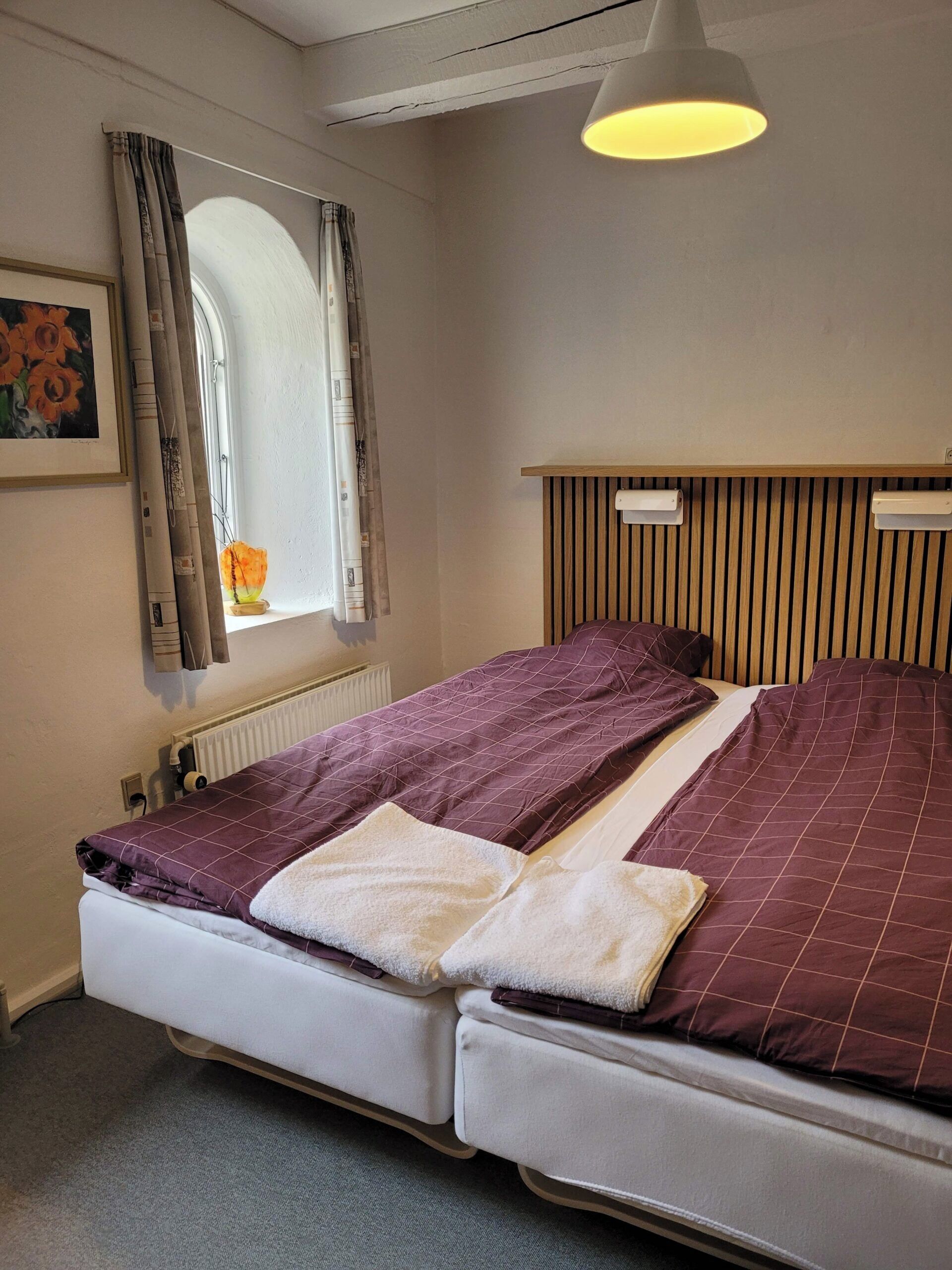 Una cama con cabecera, una ventana con cortinas, un radiador y un cuadro en la pared.
