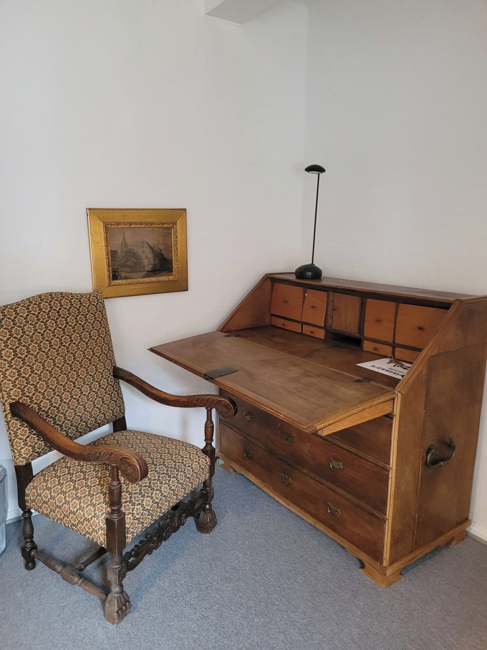 A room with a wooden desk, a chair, a lamp, and a framed picture on the wall.