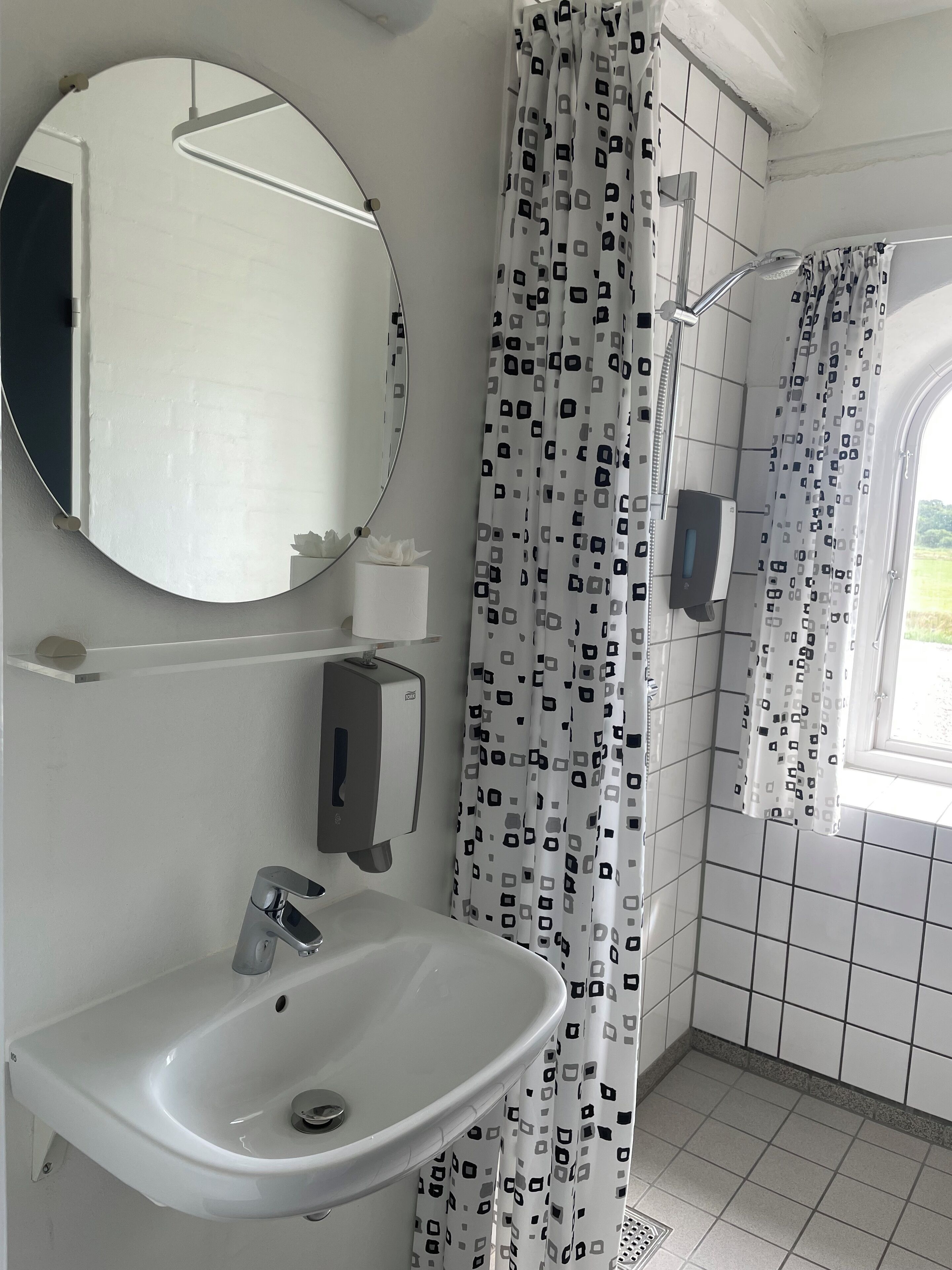 Een badkamer met een wit wastafelmeubel, een spiegel, een douchegordijn met een geometrisch patroon en een raam met uitzicht op groen.