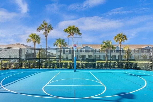 An outdoor basketball court with a blue surface, surrounded by a chain-link fence, palm trees, and a building in the background.
