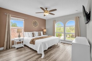 Relax in this serene bedroom with a large bed, ceiling fan, and flat-screen TV, offering a peaceful view of the surrounding greenery.