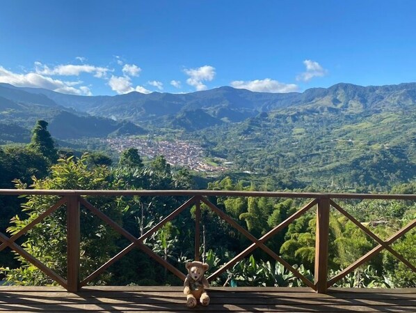 A wooden deck with a teddy bear, overlooking a mountainous landscape with a town below. - Finca Milena Jardin Antioquia (Jardin)