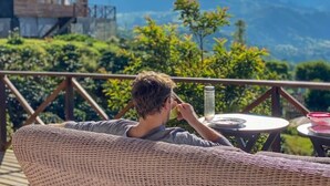 Una persona sentada en una silla de mimbre contemplando un paisaje montañoso, con un edificio de madera al fondo.