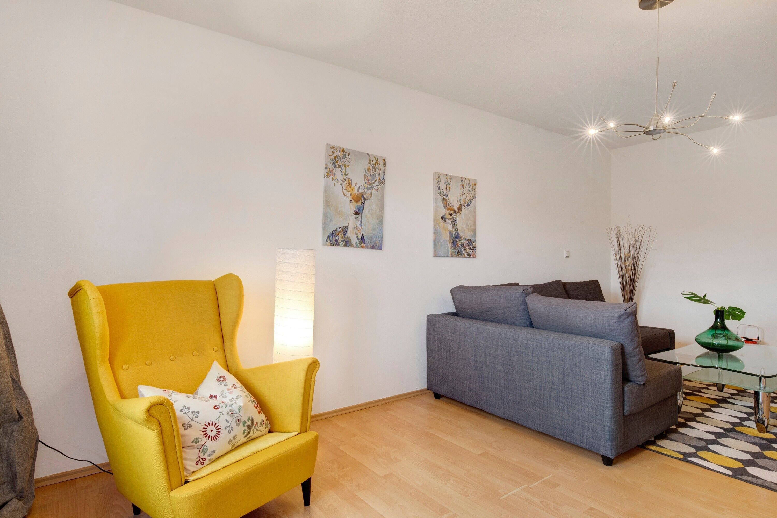 Een woonkamer met een gele fauteuil, een grijze bank, twee aan de muur bevestigde schilderijen, een moderne kroonluchter en een kamerkleed met dessin.