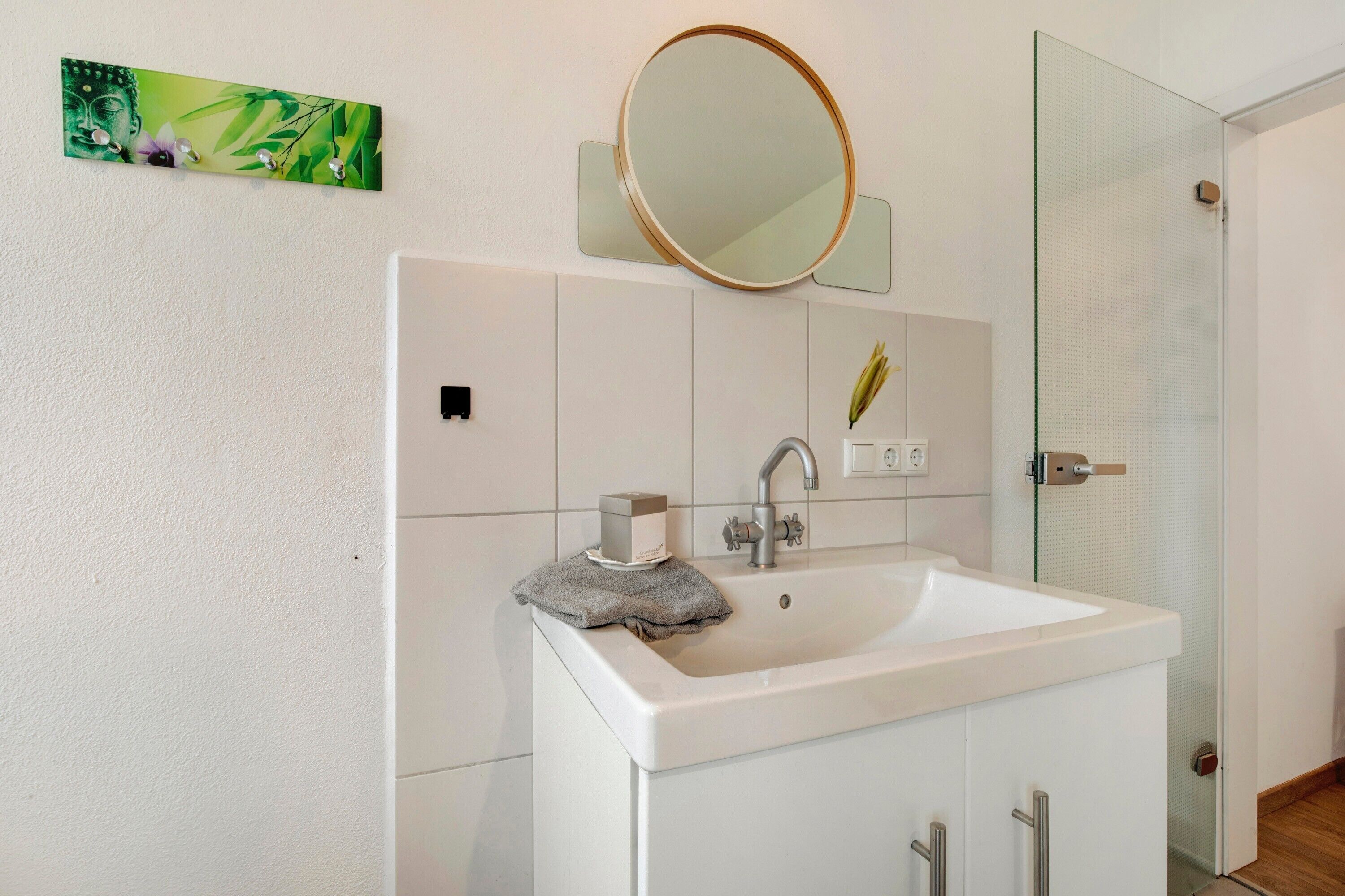 Ein Badezimmer mit einem weißen Waschbecken, einem runden Spiegel, einem Handtuch und einem grünen Dekoelement an der Wand.