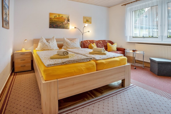 Un lit double avec une literie jaune, une table de chevet avec une lampe, un canapé rouge, une petite table avec un bol, et une fenêtre avec des rideaux.