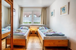 A bedroom with two wooden beds, a mirror, a window with curtains, and a small table with a lamp.