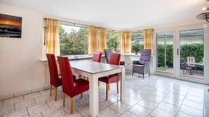 A dining area with a table and chairs, large windows with curtains, and a view of greenery outside. - Apartment 'Prerow Am Deich - Fewo 3' with Private Terrace, Private Garden and Wi-Fi (Prerow)