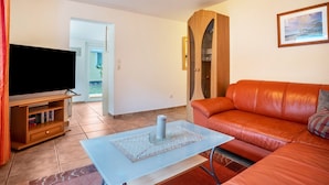 A living room with a TV, orange sofa, and a glass coffee table. - Apartment 'Prerow Am Deich - 4' with Private Terrace, Private Garden and Wi-Fi (Ostseebad Prerow)
