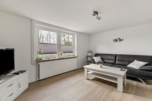 A modern living room with a black leather sofa, a white coffee table, a television on a white cabinet, and a window with blinds.