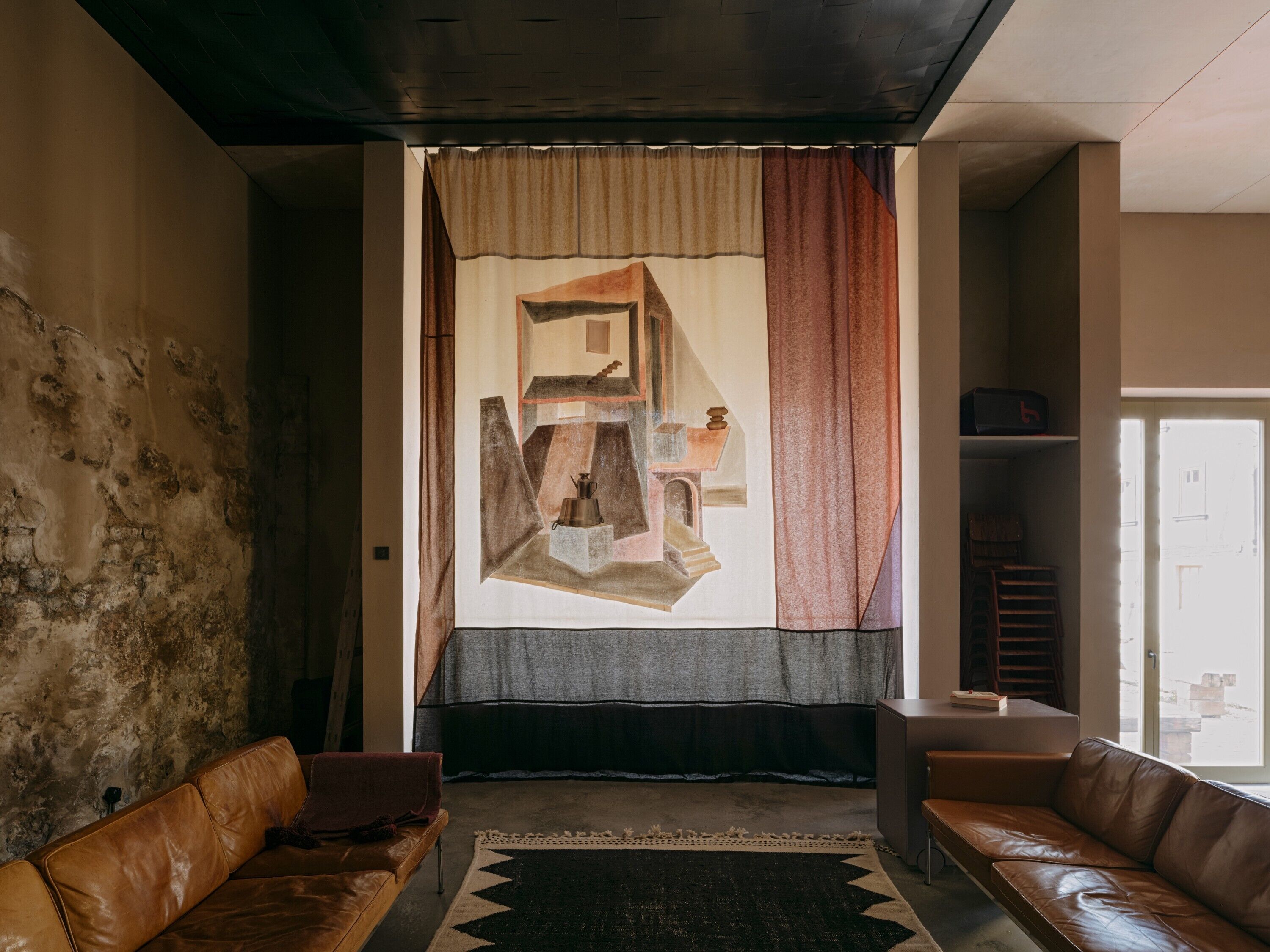 Een kamer met een groot raam, twee leren banken, een kamerkleed met een patroon en een geometrisch schilderij aan de muur.