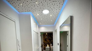 A hallway with white walls, a marble floor, a ceiling with perforated panels, and a mirror on the right wall.