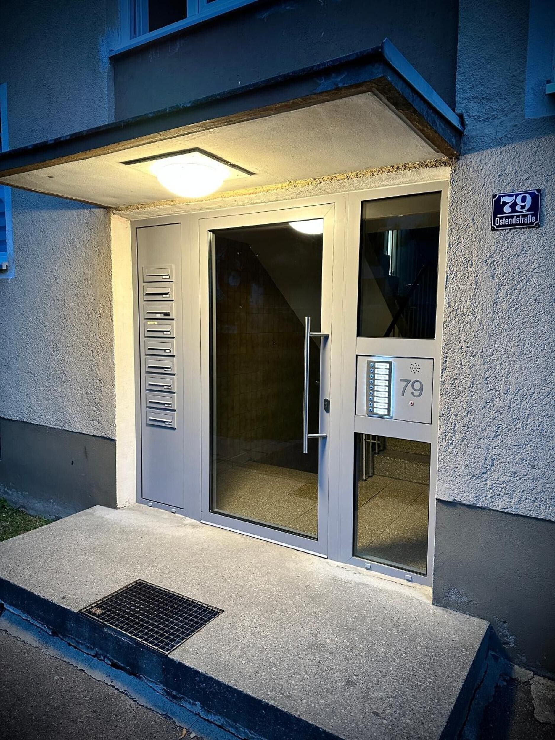 A building entrance with a glass door, a metal mail slot, and a sign indicating the address '79 Ostendstrasse.'