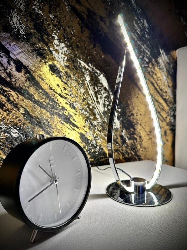 A modern alarm clock with a metallic base and a curved light bar, placed on a white surface against a textured wall.