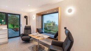 Une pièce moderne comprenant une table à manger, deux chaises, un canapé et de grandes fenêtres donnant sur un paysage verdoyant.