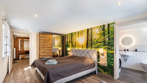 A modern bedroom with a large bed, a built-in wardrobe, a bathroom with a mirror and sink, and a forest-themed wall mural. - Apartment 'Ferienwohnung Am Augusttal' with Mountain View, Private Terrace and Wi-Fi (Hainewalde)