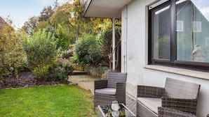 Eine Terrasse mit Korbmöbeln, einem Glastisch und Blick auf einen Garten mit Bäumen und einem Gebäude im Hintergrund.