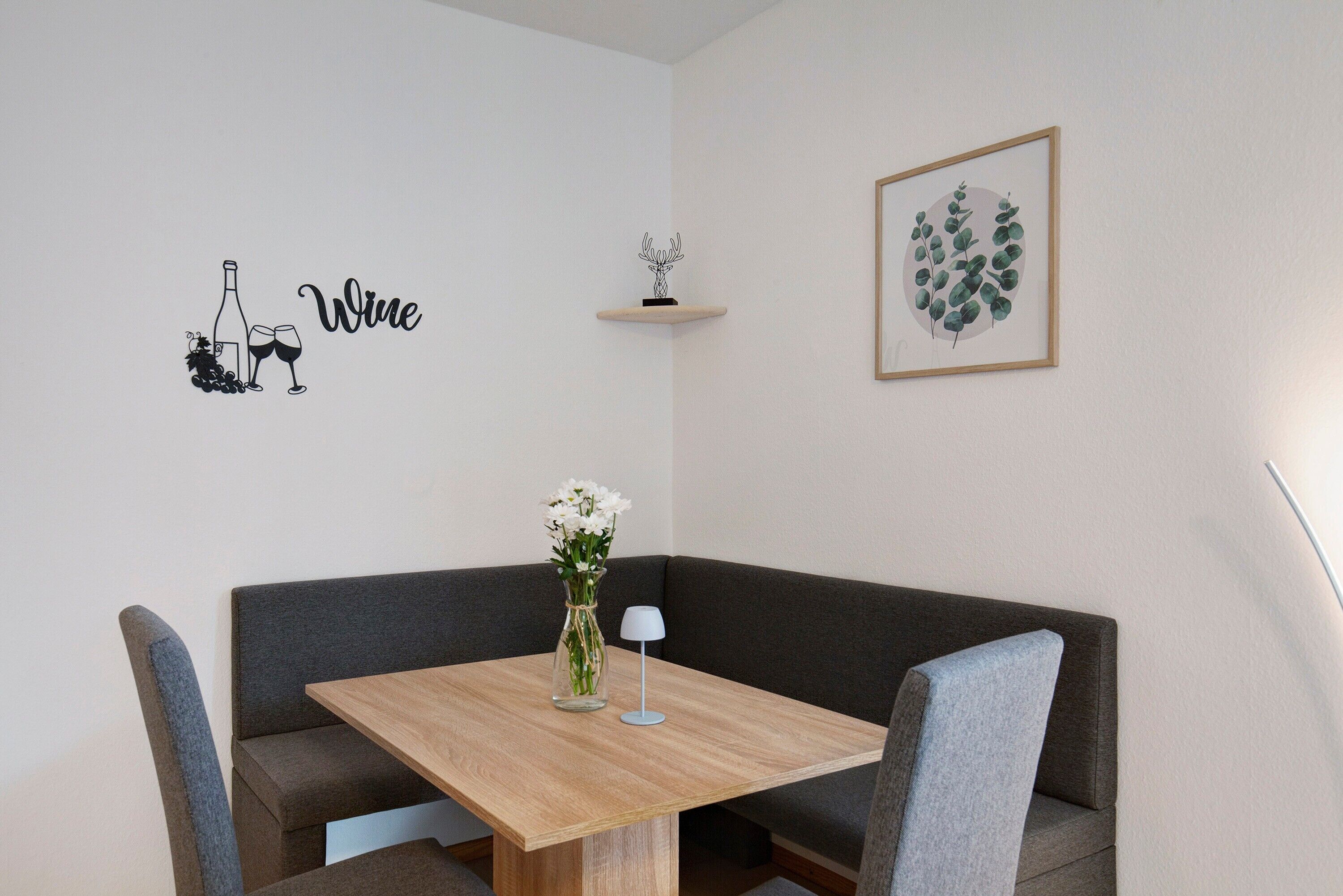 Un'area pranzo angolare con un tavolo in legno, sedie grigie rivestite in tessuto, un vaso con fiori, una piccola lampada e opere d'arte tra cui un'illustrazione di vino, una mensola con un oggetto decorativo e una pianta in cornice.