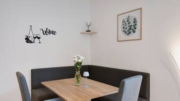 Un'area pranzo angolare con un tavolo in legno, sedie grigie rivestite in tessuto, un vaso con fiori, una piccola lampada e opere d'arte tra cui un'illustrazione di vino, una mensola con un oggetto decorativo e una pianta in cornice.