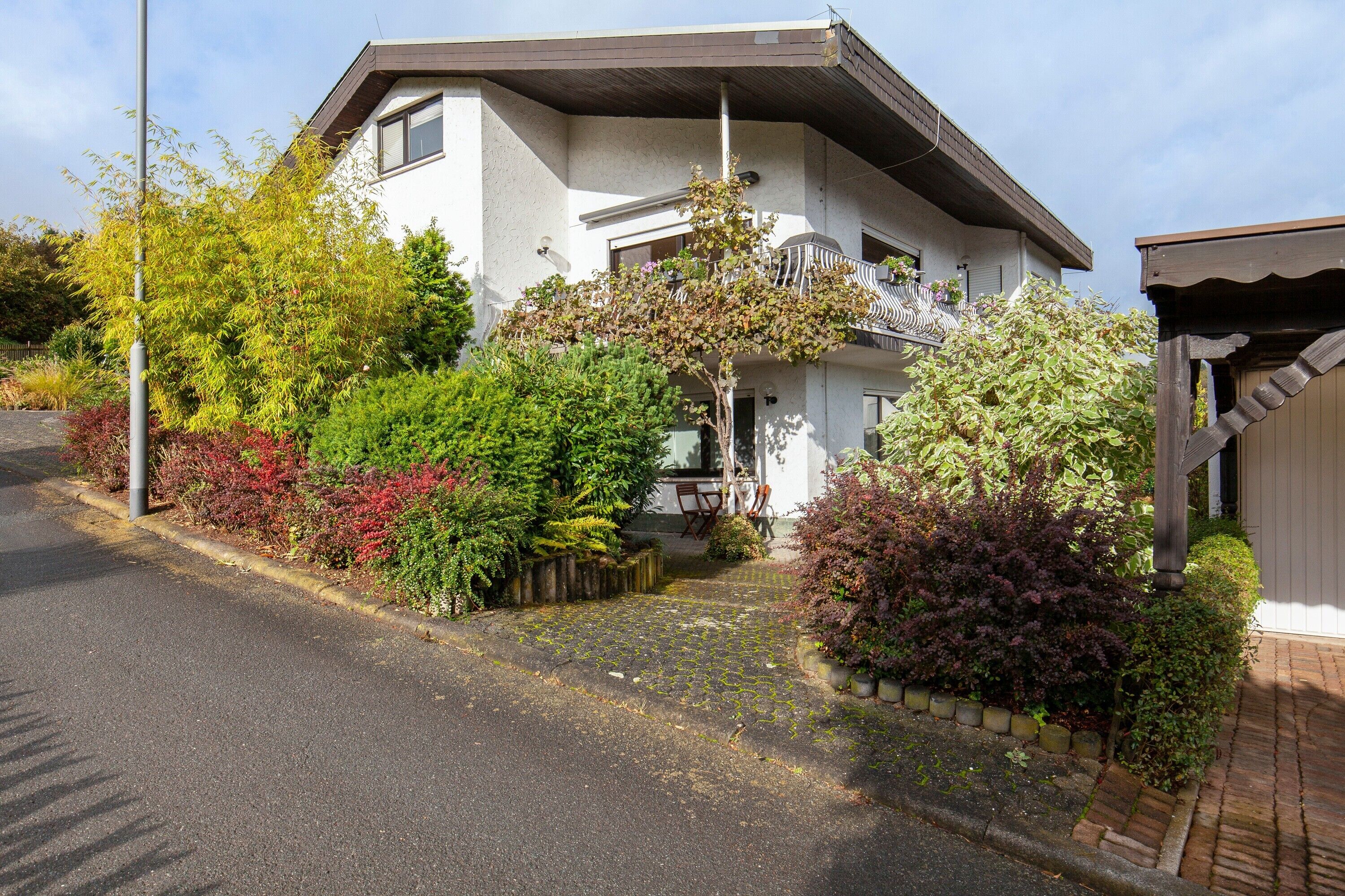 Ein zweistöckiges Haus mit überdachter Veranda, einer sichtbaren Treppe und einem Garten mit verschiedenen Pflanzen.