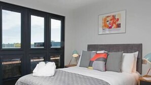 A modern bedroom with a large bed, a headboard, a bedside table with a lamp, and a window with a view.