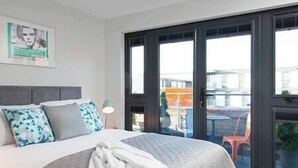 A hotel room with a large bed, a view of the city through the windows, and a balcony with a table and chairs.