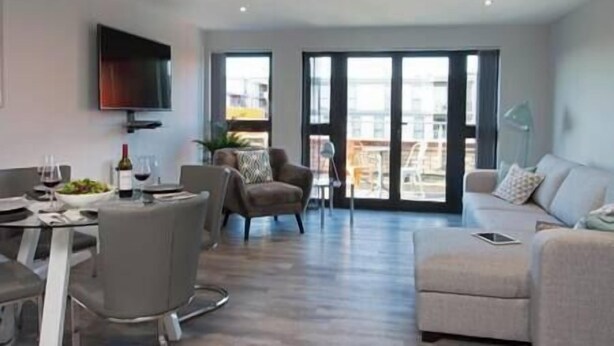 A modern living room with a dining area, a flat-screen TV mounted on the wall, and a balcony with a view.