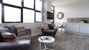 Una sala de estar moderna con un sofá gris, una mesa de centro redonda y un sillón negro. Se observa una zona de cocina con encimera gris y un espejo redondo en la pared.