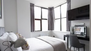 A modern bedroom with a bed, a desk, and a chair. There are large windows with curtains, a flat-screen TV, and a radiator.