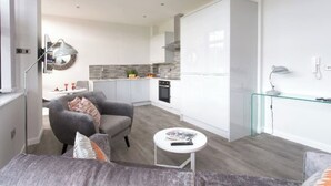 A modern apartment with a grey sofa, a small round table, and a kitchen with white cabinets and a grey backsplash.
