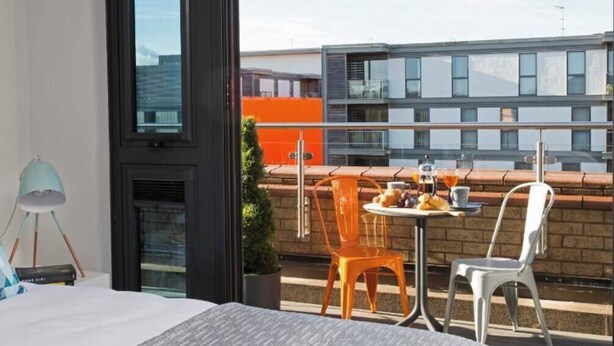 A modern hotel room with a balcony, a dining table with chairs, and a view of the cityscape.