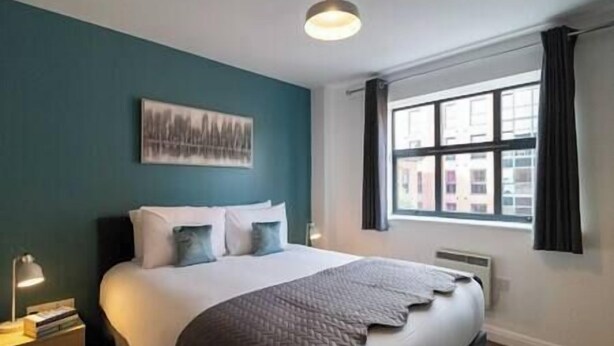 A hotel room with a large bed, a nightstand, a desk lamp, a window with curtains, and a cityscape view.