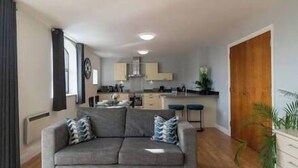 A modern apartment with a grey sofa, a kitchen with wooden cabinets, and a wooden door.