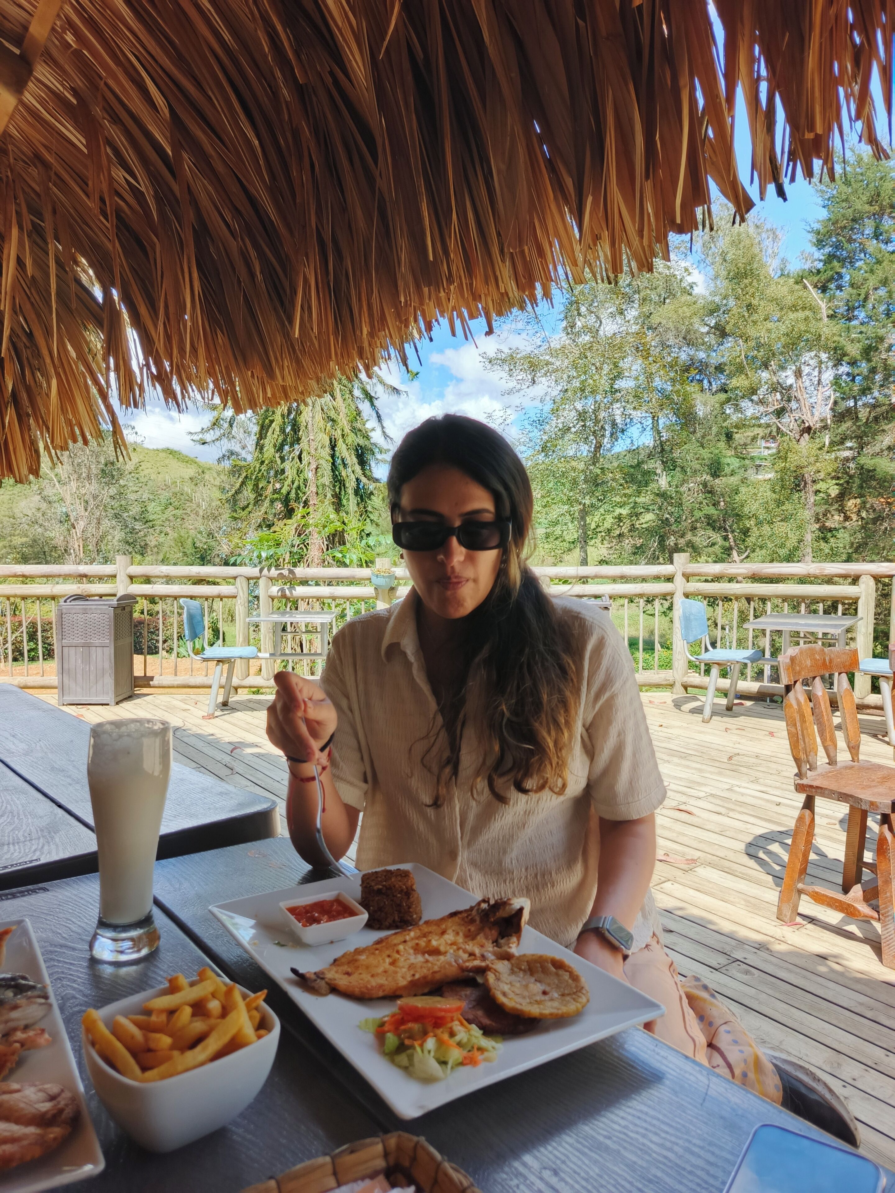 A person dining at an outdoor table with a thatched roof, surrounded by greenery and a wooden deck.