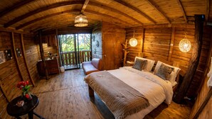 A wooden cabin bedroom with a bed, a bench, a table with a vase, and a view through a door. - Refugio Natural - Guarne - Cabañas Ecológicas con Jacuzzi e hidromasaje (Guarne)