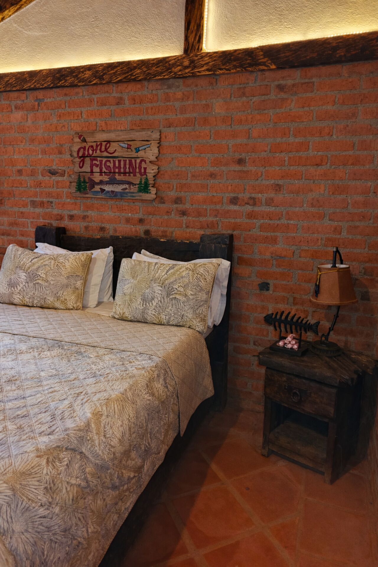 A bedroom with a brick wall, a bed with patterned bedding, a nightstand with a lamp, and a sign that says 'Gone Fishing'.