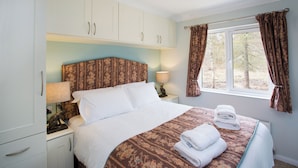 A neatly made bed with a patterned bedspread and folded towels, a nightstand with a lamp, and a window with curtains. - Bluebell Lodge - Sleeps 4,hot Tub, Loch & Woodland (Strathpeffer)