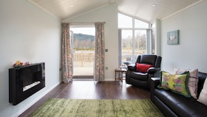 A living room with a fireplace, a leather sofa, a coffee table, and a view of the outdoors. - Bluebell Lodge - Sleeps 4,hot Tub, Loch & Woodland (Strathpeffer)