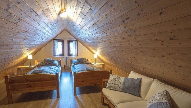 Une chambre confortable située sous les toits, avec deux lits en bois, un canapé et une fenêtre laissant entrer la lumière naturelle.