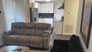 Una sala de estar con un sofá seccional gris, un sofá negro y una mesa de centro. Se observa un área de cocina al fondo, con gabinetes blancos y un salpicadero negro.