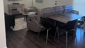 Una sala moderna con un sofá seccional, una mesa de comedor con sillas, un televisor de pantalla plana, un ventilador de techo y una lámpara de pie.