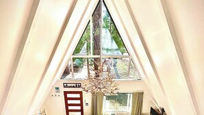 A loft-style room with a sloped ceiling, a large window, a green sofa, a wooden dining table, and a television.