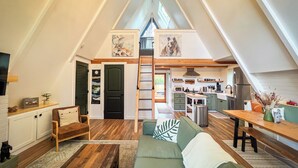 A cozy loft apartment with a high ceiling, a dining area, a kitchen, and a living room with a green sofa and wooden flooring.