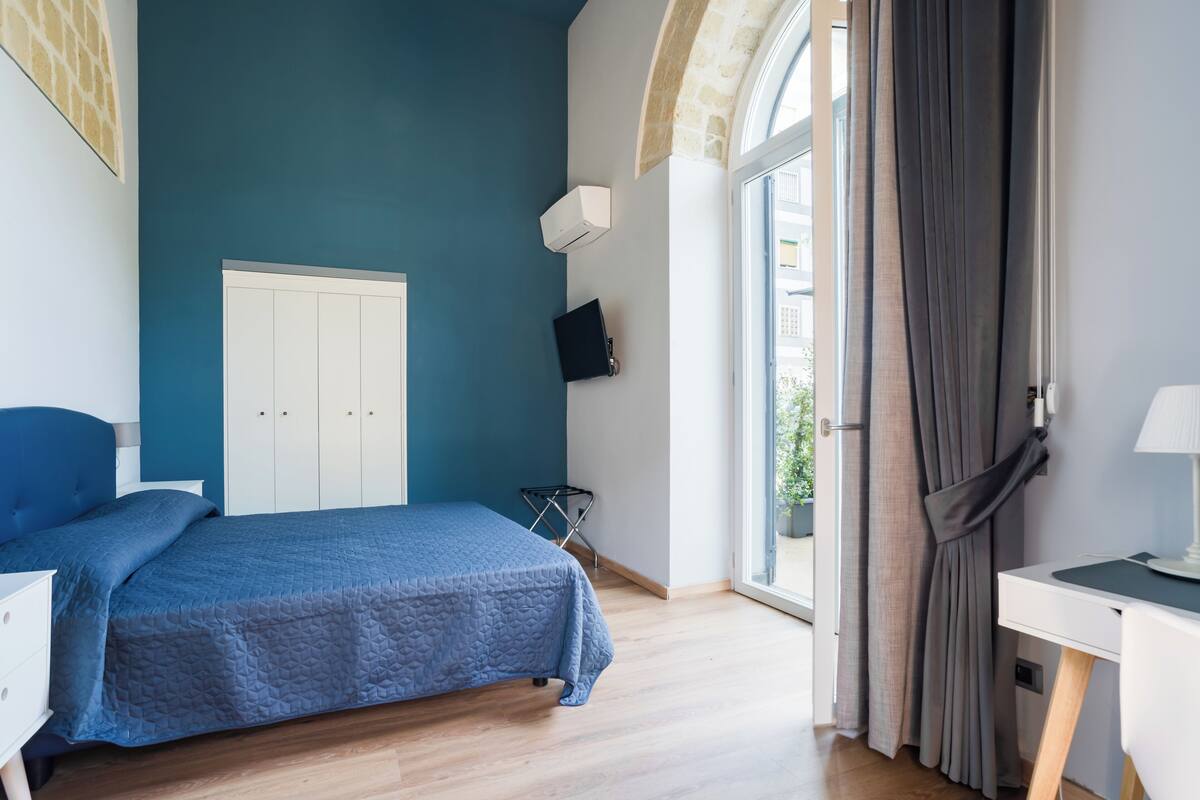 A bedroom with a bed, a desk, a chair, a wardrobe, and a window with curtains.