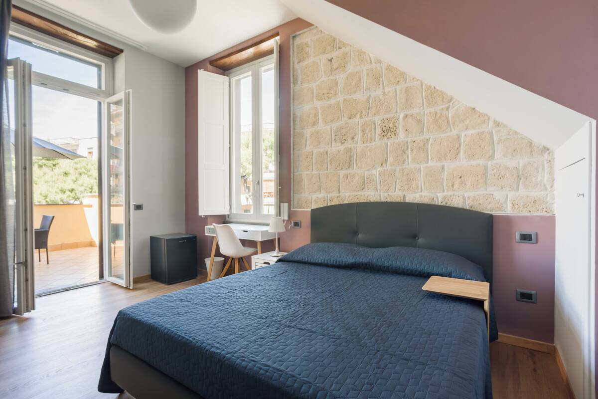 A bedroom with a large bed, a desk, and a chair, a stone wall, and a sliding glass door to an outdoor area.