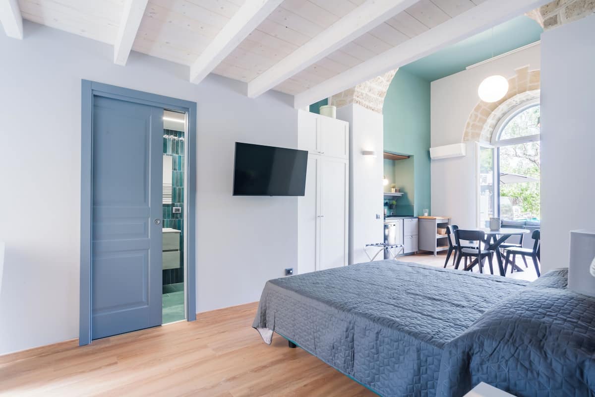A bedroom with a bed, a TV, and a view of a kitchen and dining area.