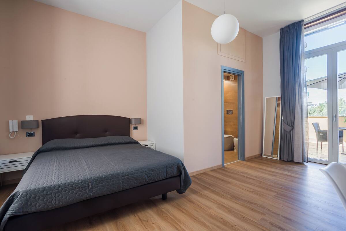 A modern bedroom with a large bed, bedside tables, a wall-mounted lamp, and a sliding glass door leading to a balcony.
